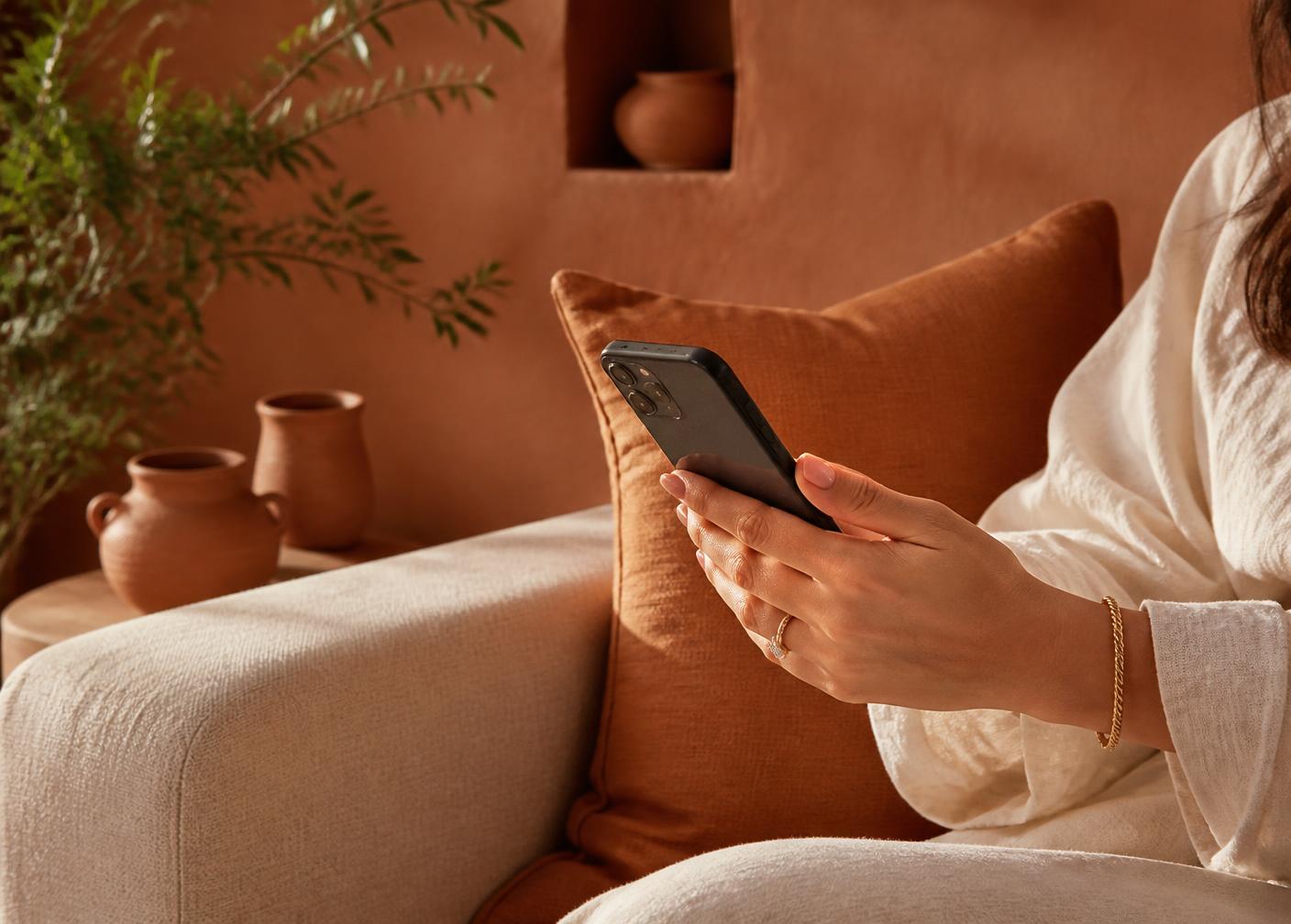 Mulher sentada em um sofá claro, com celular nas mãos, em uma sala de paredes terracota